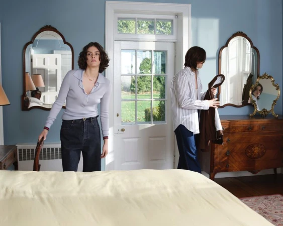 Two adult women in an indoor bedroom setting with a bed and antique furniture, natural light, and mirrors reflecting the scene.