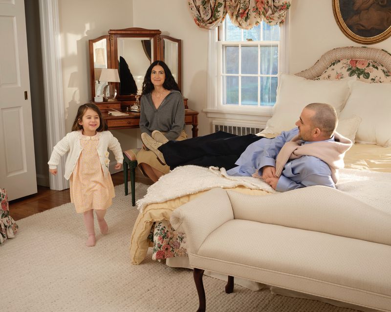 Family scene in a bedroom: A young girl walks toward the camera, a woman sits on a bench, and a man reclines on a bed in soft lighting.