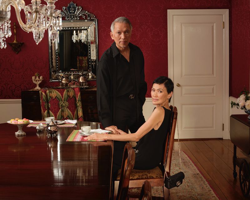 Elegant couple poses in dining room with chandelier, mahogany table set with tea service, and dark red wallpaper with ornate mirror.