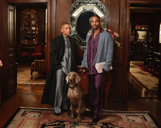 Two adult males pose with a Weimaraner dog inside a wood-paneled room featuring ornate architectural details, a patterned rug, and doors.