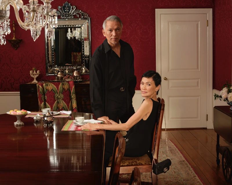 An older man and woman in black clothing posed in an opulent dining room with red patterned wallpaper for the Gucci Together 2025 campaign.