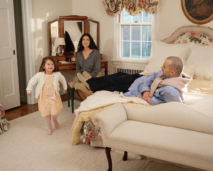A young girl running in a bedroom while a man rests on a bed and a woman sits nearby at a vanity table.