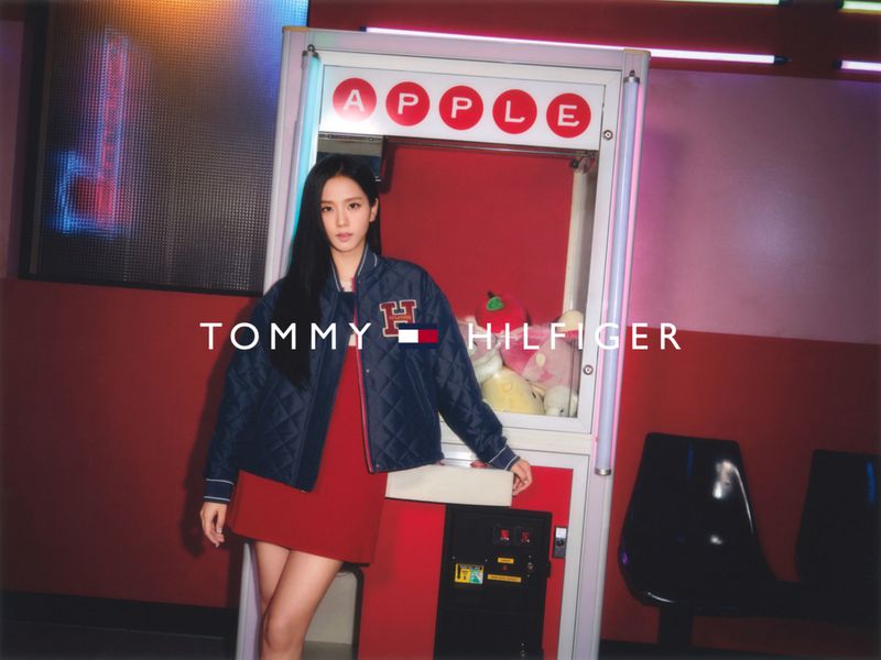 A young woman in a red dress and blue Tommy Hilfiger jacket stands in front of a claw machine in an indoor setting.