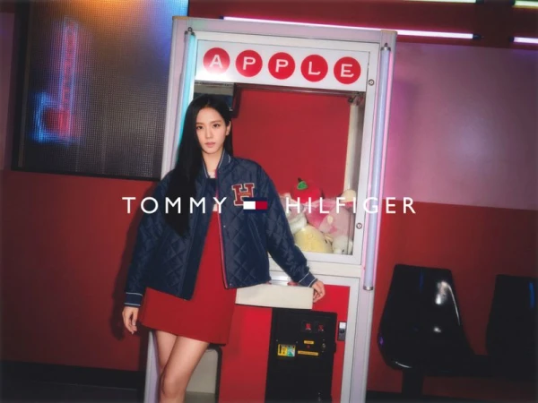 A young woman in a red dress and blue Tommy Hilfiger jacket stands in front of a claw machine in an indoor setting.