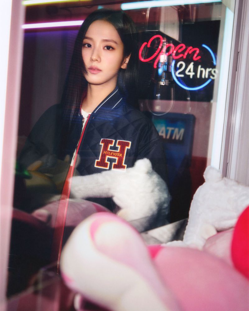 A stylish young woman with long dark hair wears a Tommy Hilfiger jacket in a neon-lit indoor setting with a playful aesthetic.