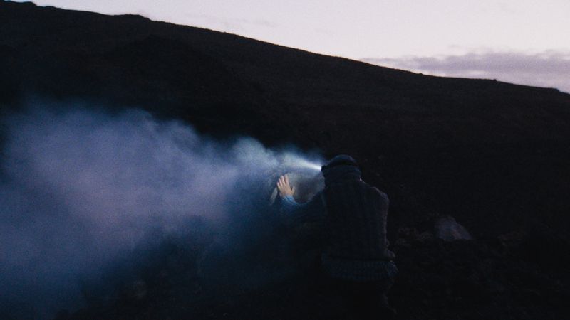 Person with headlamp kneels on a hillside at dusk. The light illuminates smoke or fog in front with a dark, mountainous background.