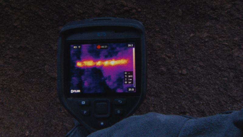 A handheld thermal imaging camera displays a heat signature against a dark background, showing temperature variations with a purple hue.