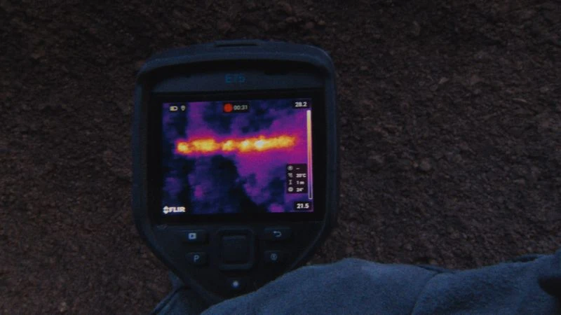 A handheld thermal imaging camera displays a heat signature against a dark background, showing temperature variations with a purple hue.