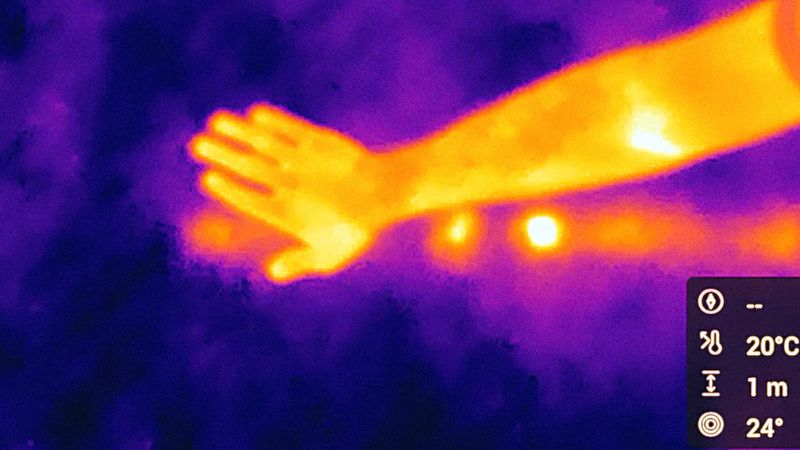 Thermal imaging shows a human arm and hand in vibrant oranges and yellows, set against a cool, contrasting purple background, displaying temperature variations.
