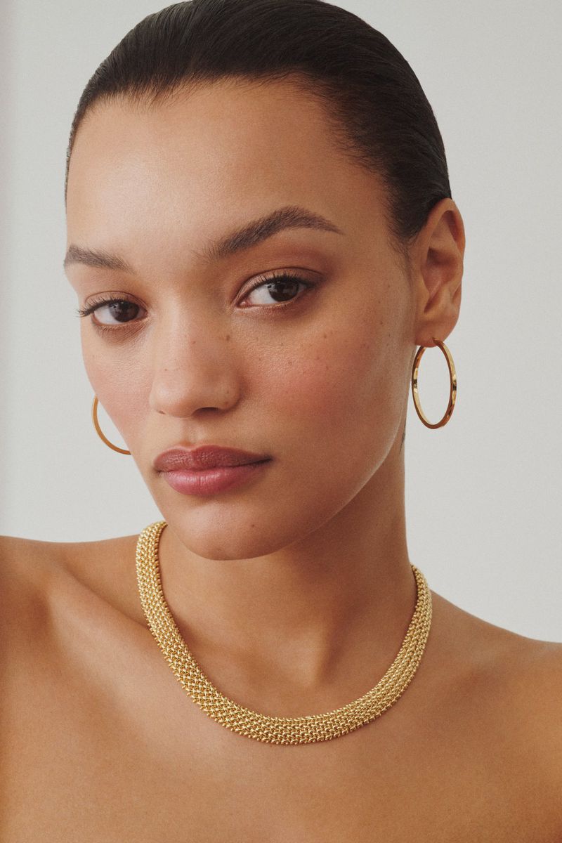 Close-up portrait of model Lameka Fox wearing a textured gold necklace and hoop earrings against a neutral background.