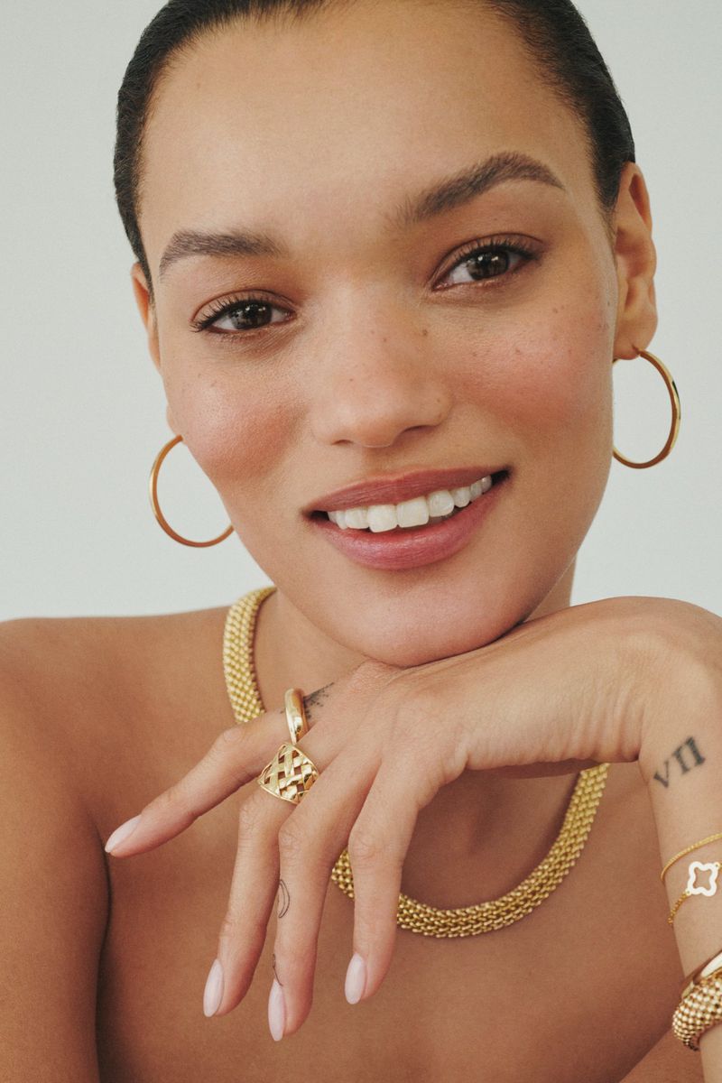 Close-up portrait of model Lameka Fox wearing gold hoop earrings and fine jewelry necklaces against a neutral background