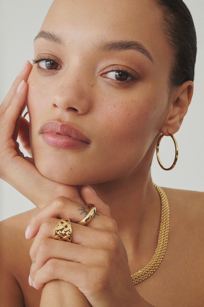 Close-up portrait of model Lameka Fox wearing gold hoop earrings, chunky rings, and a textured gold chain necklace against a white background.