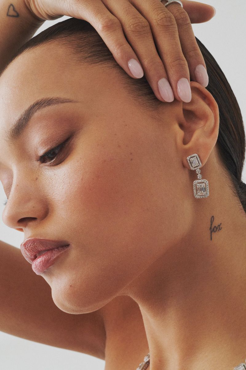 Close-up studio shot of a fair-skinned woman wearing diamond earrings and a ring, nails painted light pink, against a white background.