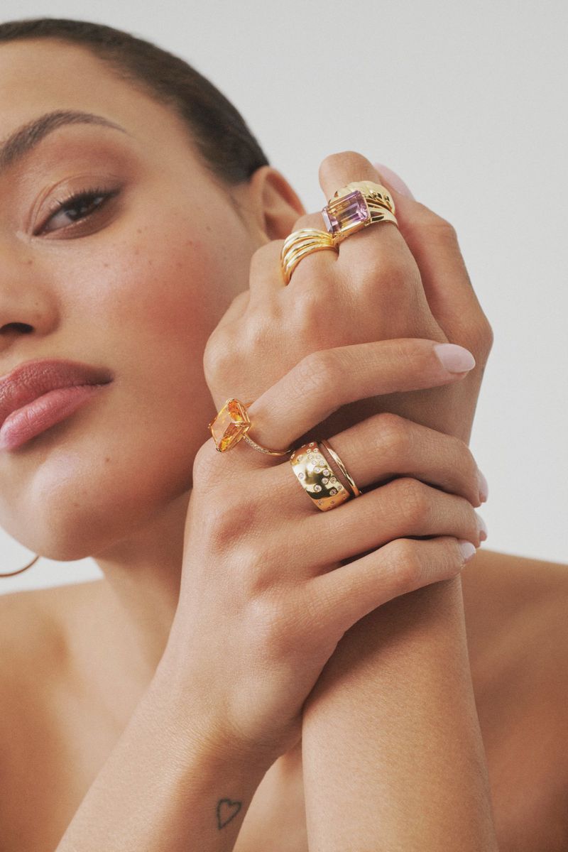 Close-up of a fair-skinned adult female's hands adorned with yellow gold rings featuring various gemstone settings, against a plain backdrop.