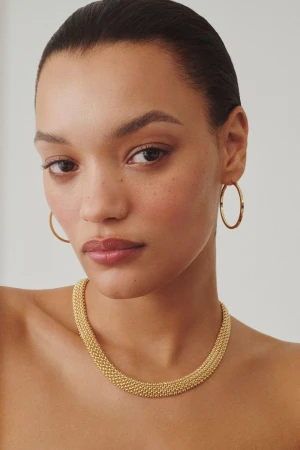 Close-up portrait of model Lameka Fox wearing a textured gold necklace and hoop earrings against a neutral background.