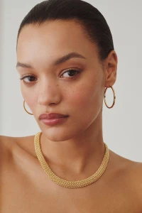 Close-up portrait of model Lameka Fox wearing a textured gold necklace and hoop earrings against a neutral background.