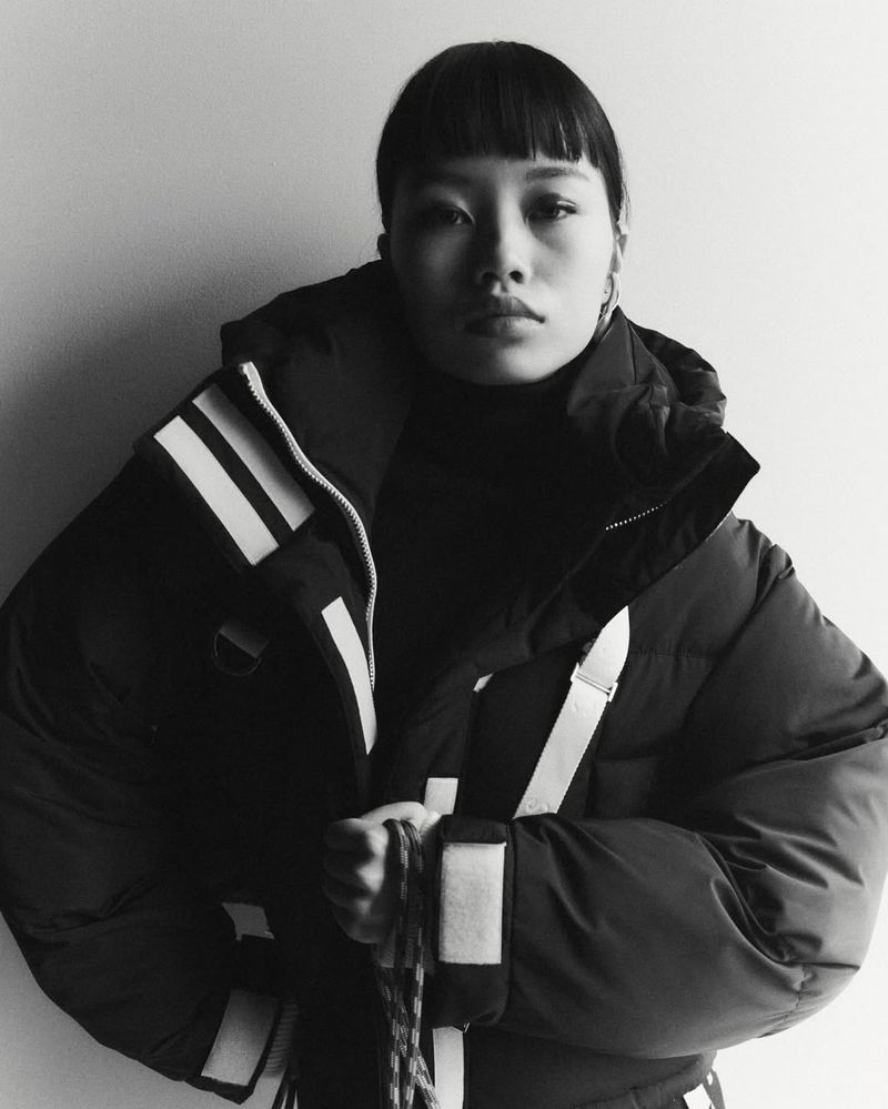 Monochrome portrait of Asian woman in black puffer jacket with white stripes, looking directly at the camera in a neutral indoor setting.