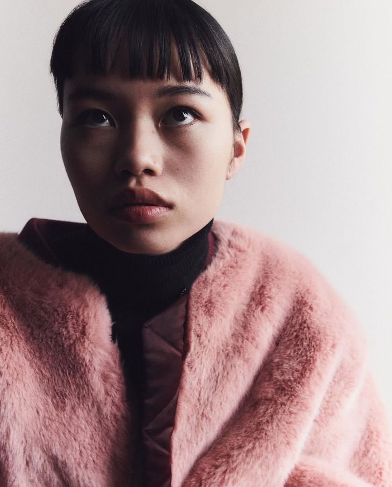 Portrait of a young adult Asian woman with bangs looking upward, wearing a black turtleneck and fuzzy pink jacket in a bright indoor setting.
