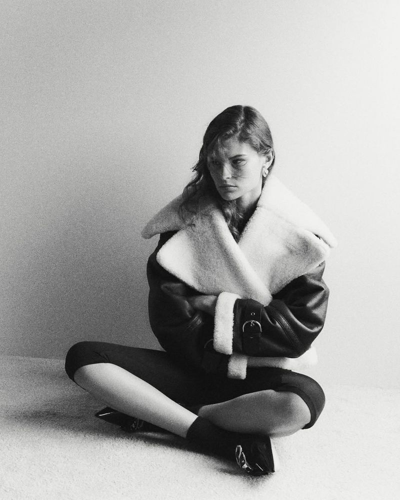 Model seated cross-legged wearing a heavy shearling jacket against a neutral studio background