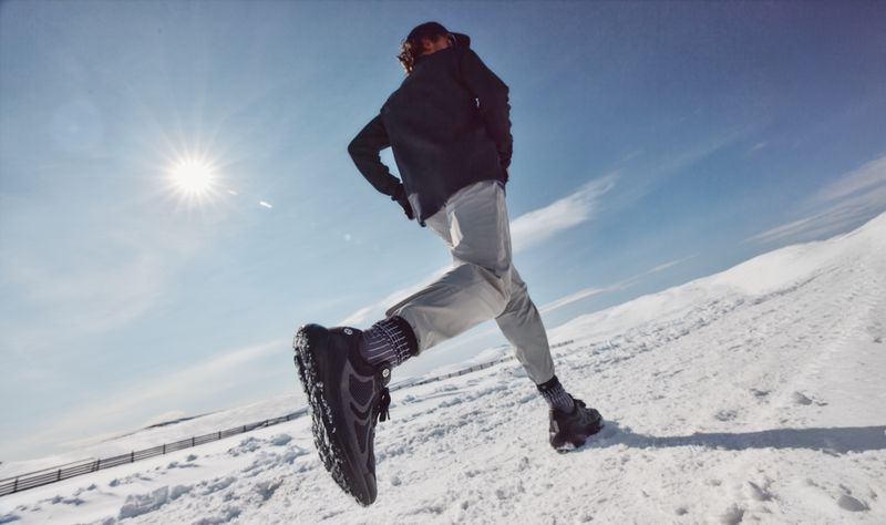 Runner in dark jacket and light trousers running uphill across a snowy landscape under a bright sun