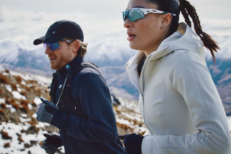 Athletic couple running outdoors on a rocky, snow-dusted mountain terrain with distant peaks, both wearing sunglasses and sporty attire.