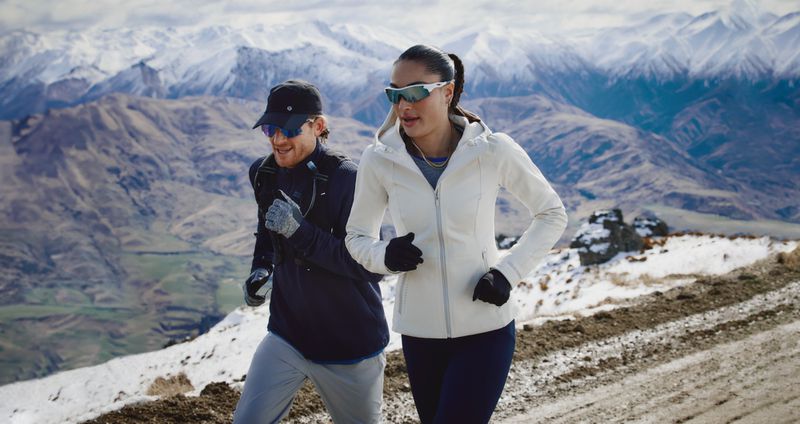 Woman and man trail running on a snowy mountain path wearing athletic clothing and sunglasses with a scenic mountain landscape in background.