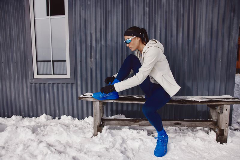 A woman sits on a wooden bench in the snow, tying the laces of a bright blue running shoe. She wears athletic wear and mirrored sunglasses.
