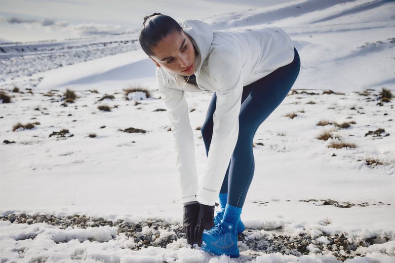 A female runner bends forward to stretch her legs on a snowy path in a mountainous region, wearing an athletic jacket, pants, and brightly colored running shoes.