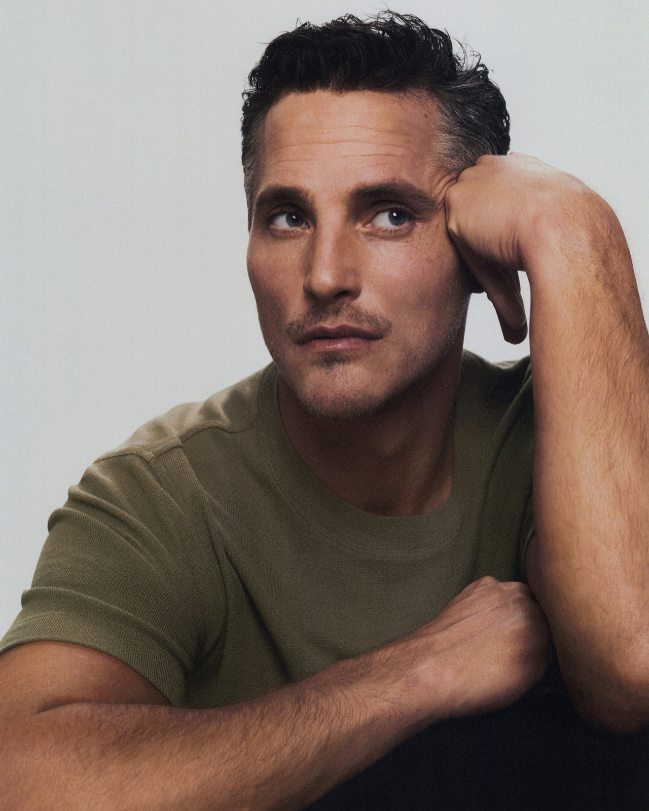 Portrait of an adult male with dark hair and a green t-shirt, posing with his hand resting on his head against a neutral background.