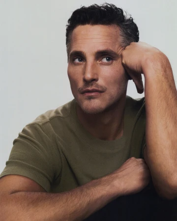Portrait of an adult male with dark hair and a green t-shirt, posing with his hand resting on his head against a neutral background.