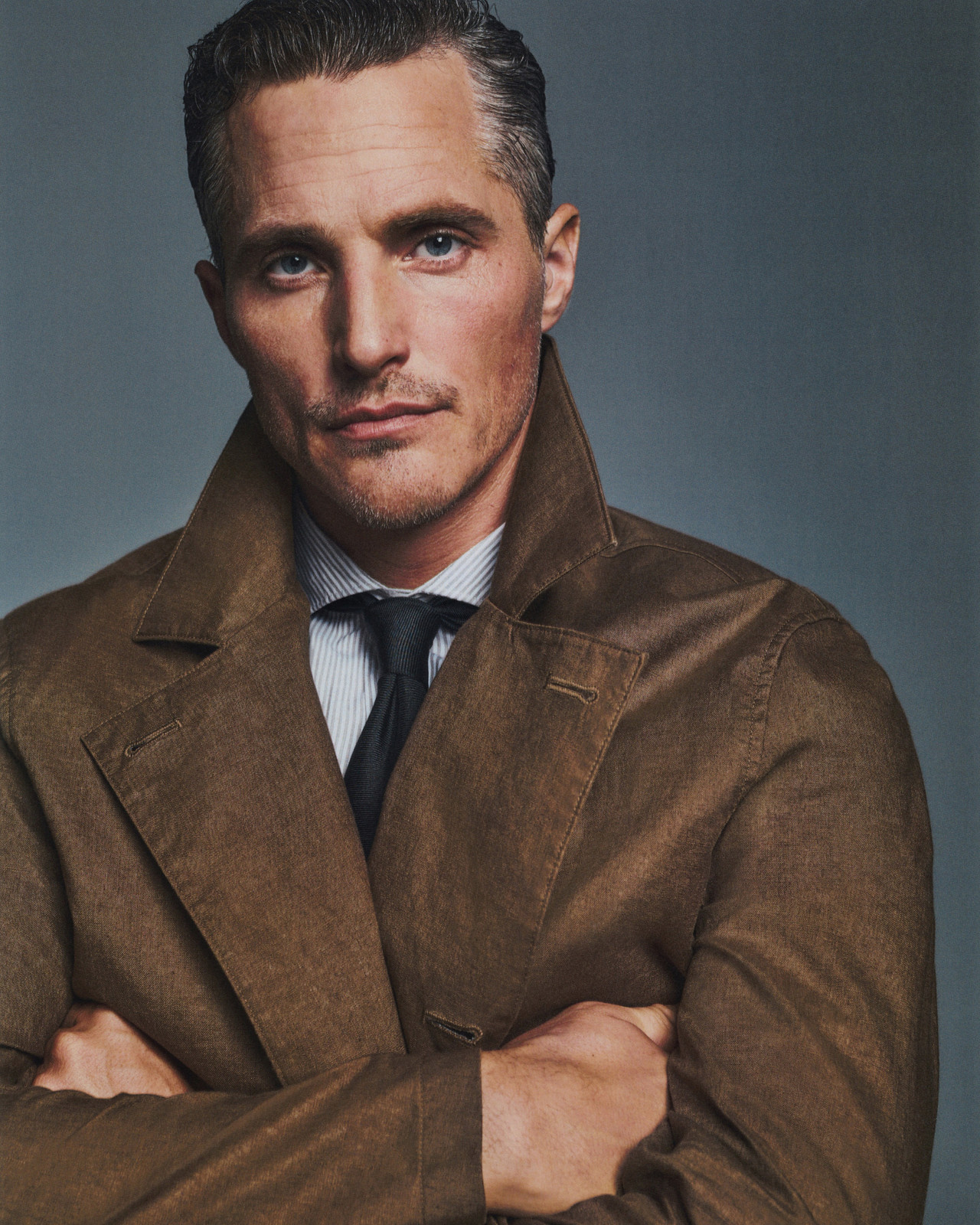 Portrait of an adult man in a brown overcoat with arms crossed, wearing a collared shirt and dark tie, set against a grey background.