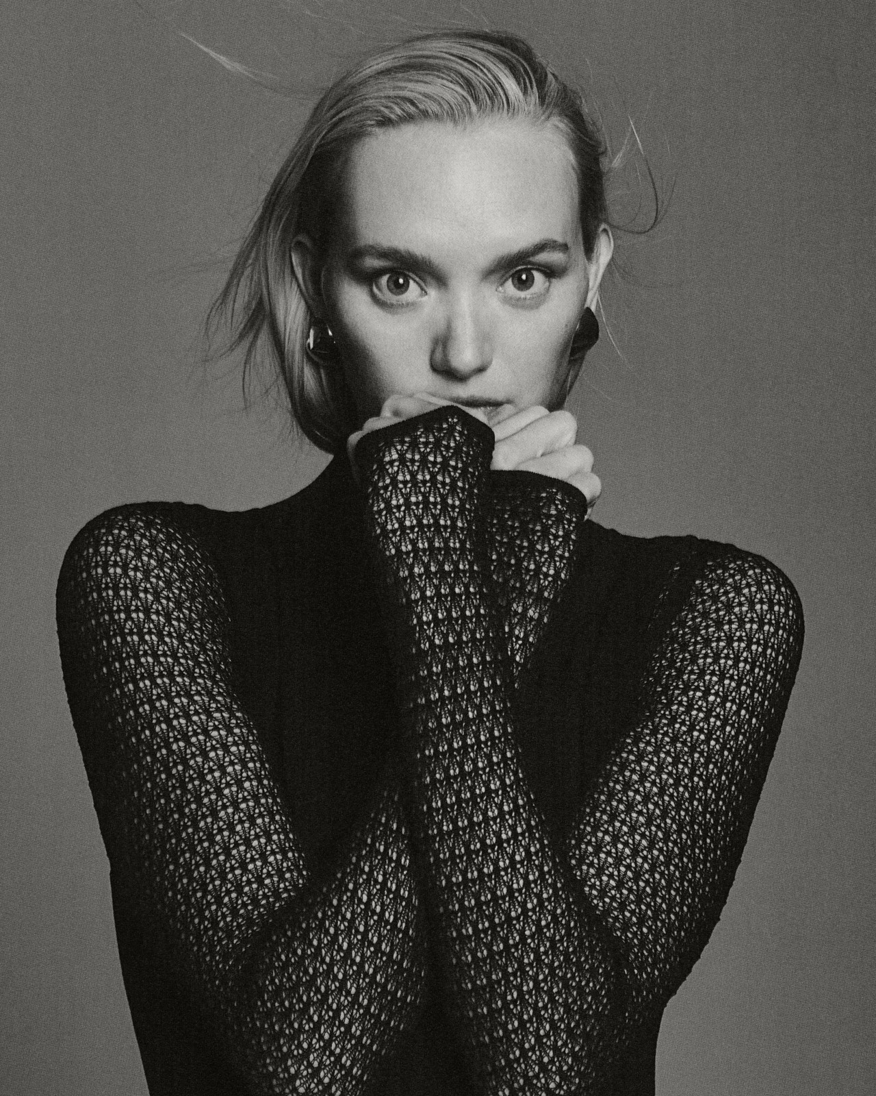 Monochrome portrait of a blonde woman with hands near her face, wearing a patterned black top and hoop earrings, set against a grey background.