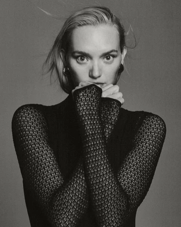 Monochrome portrait of a blonde woman with hands near her face, wearing a patterned black top and hoop earrings, set against a grey background.