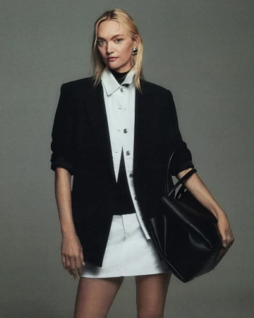 Model in black blazer and white shirt holding a black handbag against grey seamless background
