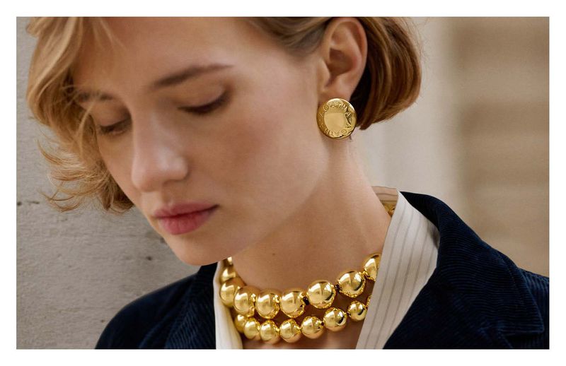 Close-up of a blonde woman wearing gold jewellery, including a ball necklace and coin earrings, set against a neutral background.