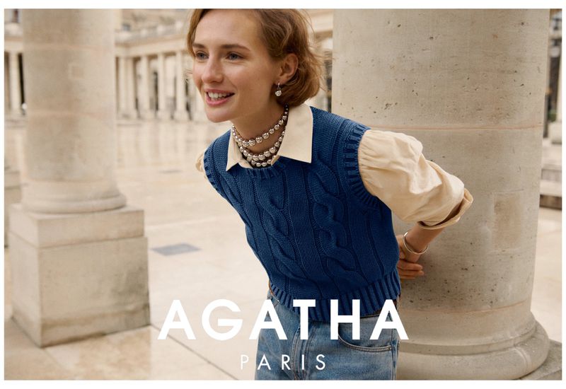 Smiling woman in a blue sweater vest and pearl necklace poses near a stone column in front of a stone building for a fashion shot.