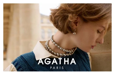 Close-up portrait of a woman wearing silver ball necklaces, a white collar shirt, and a blue knit vest against an architectural background