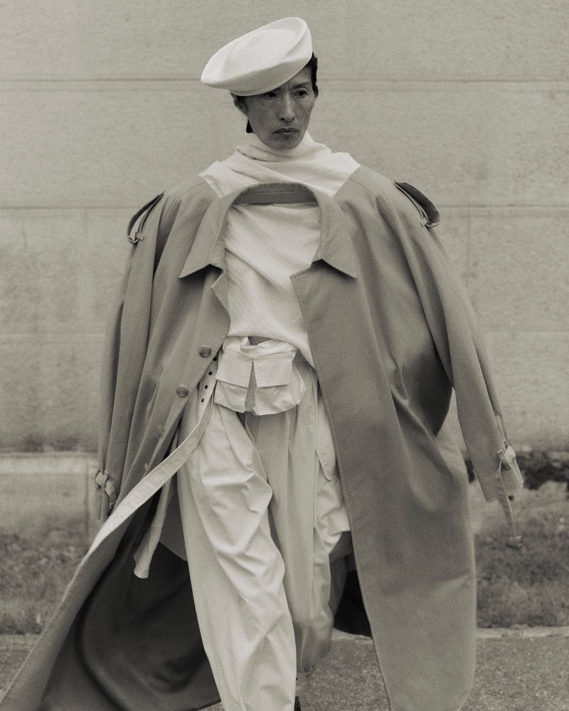 Monochromatic fashion shot of an Asian model wearing a beret, layered white top, and beige trench coat in an outdoor urban setting.