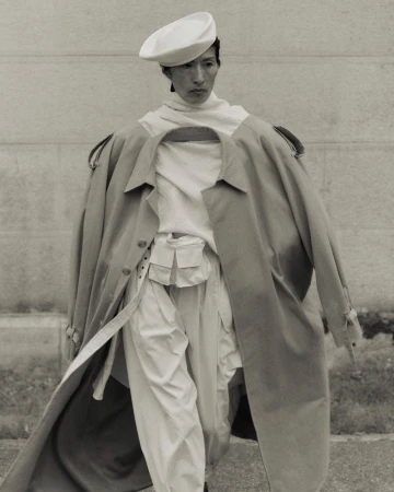 Monochromatic fashion shot of an Asian model wearing a beret, layered white top, and beige trench coat in an outdoor urban setting.