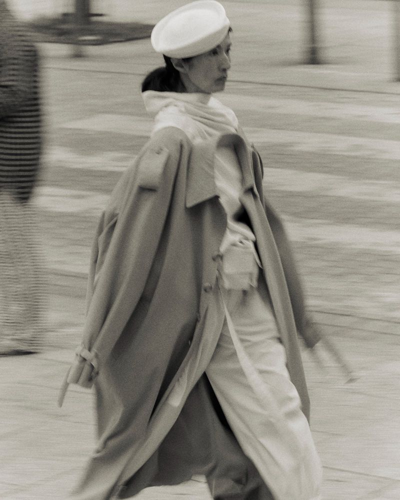 Monochrome outdoor shot of woman walking, wearing a white hat, scarf, and light-colored trench coat, with motion blur from panning.