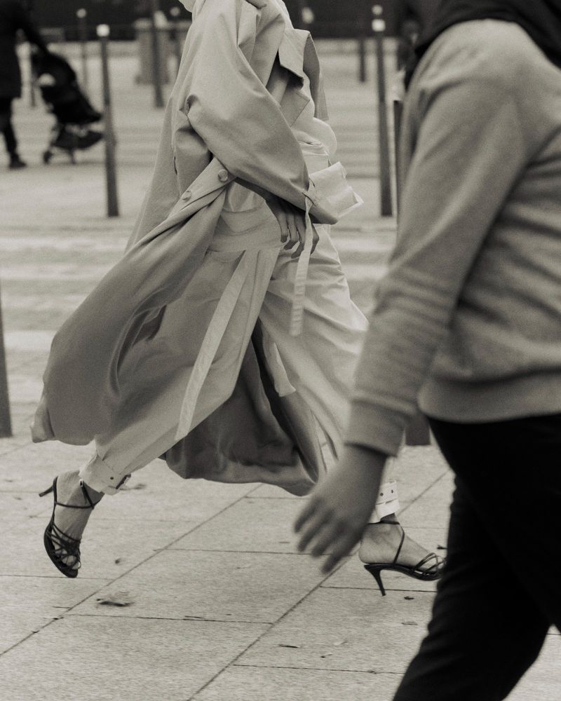 Woman strides confidently in a trench coat and heels on a city street, monochrome fashion, pedestrian in background, urban style.