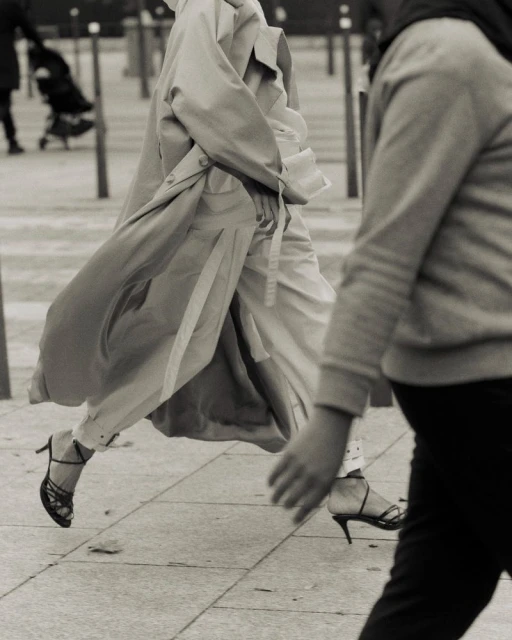 Woman strides confidently in a trench coat and heels on a city street, monochrome fashion, pedestrian in background, urban style.