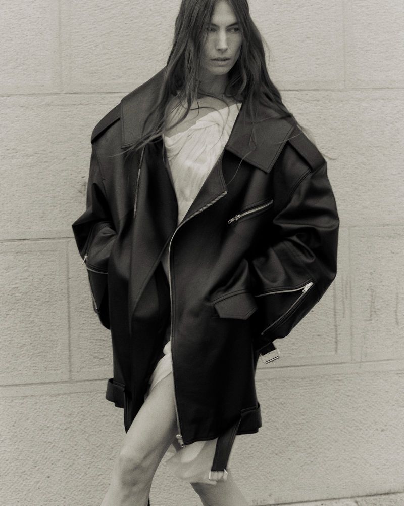 Monochrome portrait of a woman wearing an oversized black leather jacket and light-colored dress against a neutral background.