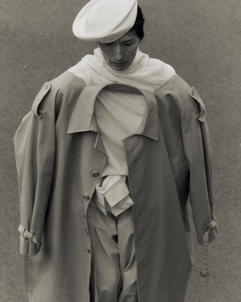 Fashion shot of an East Asian adult wearing a beige beret, white scarf, a beige trench coat over another draped around the shoulders.