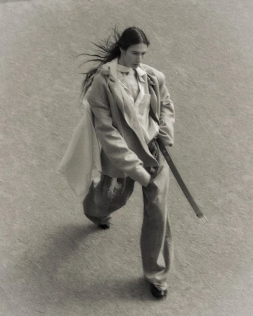 Fashionable adult strides across sandy terrain wearing a neutral blazer, pants, flowing scarf, and black dress shoes in a high-angle shot.