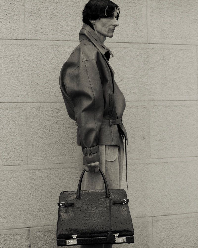 Monochrome portrait of a slender adult male in a leather jacket carrying an oversized leather briefcase against a neutral brick wall.