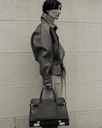 Monochrome portrait of a slender adult male in a leather jacket carrying an oversized leather briefcase against a neutral brick wall.