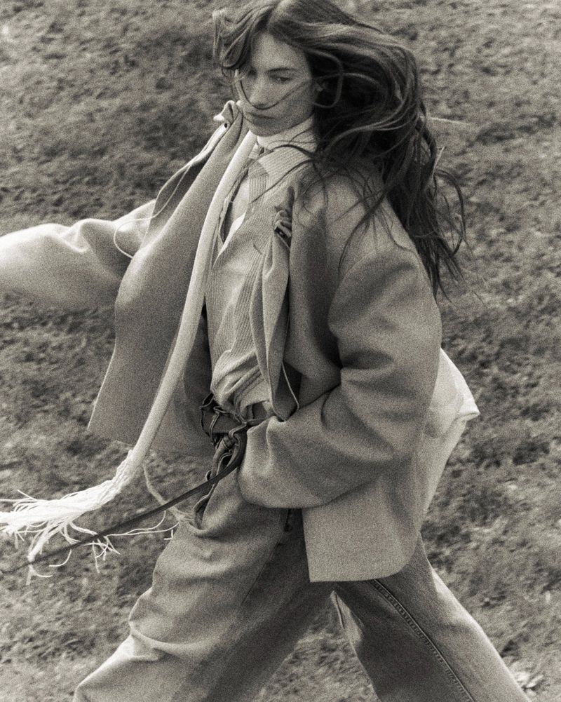 Monochrome portrait of a woman in a blazer and jeans walking through a grassy field with wind blowing her long hair.