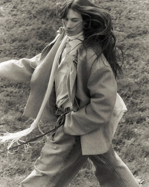 Monochrome portrait of a woman in a blazer and jeans walking through a grassy field with wind blowing her long hair.