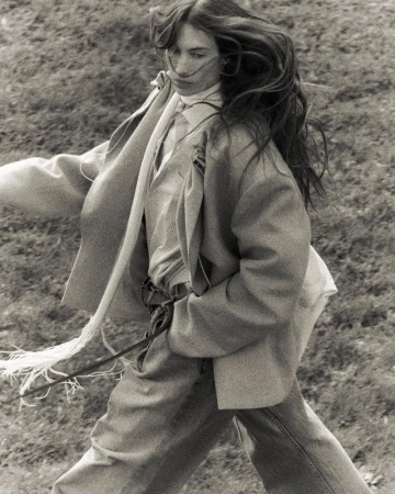 Monochrome portrait of a woman in a blazer and jeans walking through a grassy field with wind blowing her long hair.